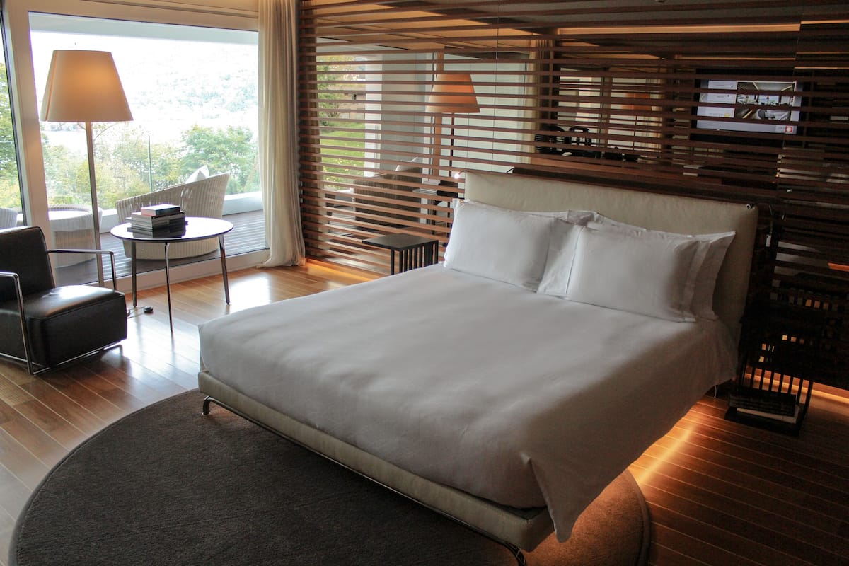 A modern hotel room with a large bed, a round rug, a small table with a lamp, and a view through large windows.