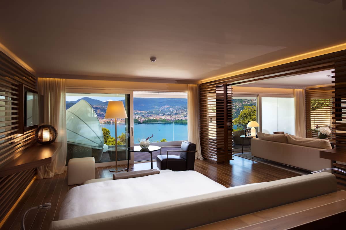 A modern hotel room with a large bed, a seating area with a table and chairs, and a balcony with a view of the city and mountains.