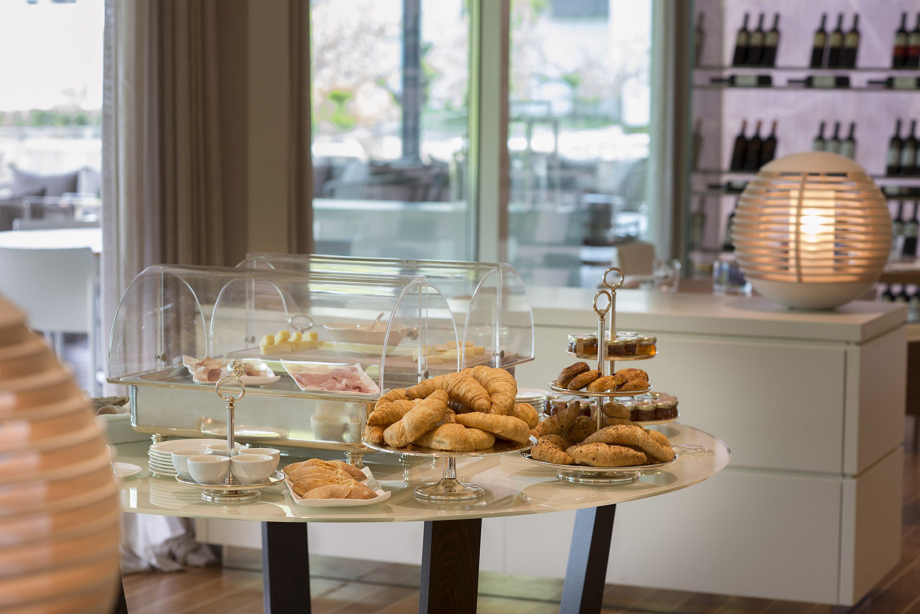 Una mesa de buffet con diversos pasteles y postres, una exhibición de vinos al fondo y una lámpara decorativa.
