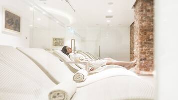A spa room with a person lying on a white bed, a brick wall, and artwork on the wall.