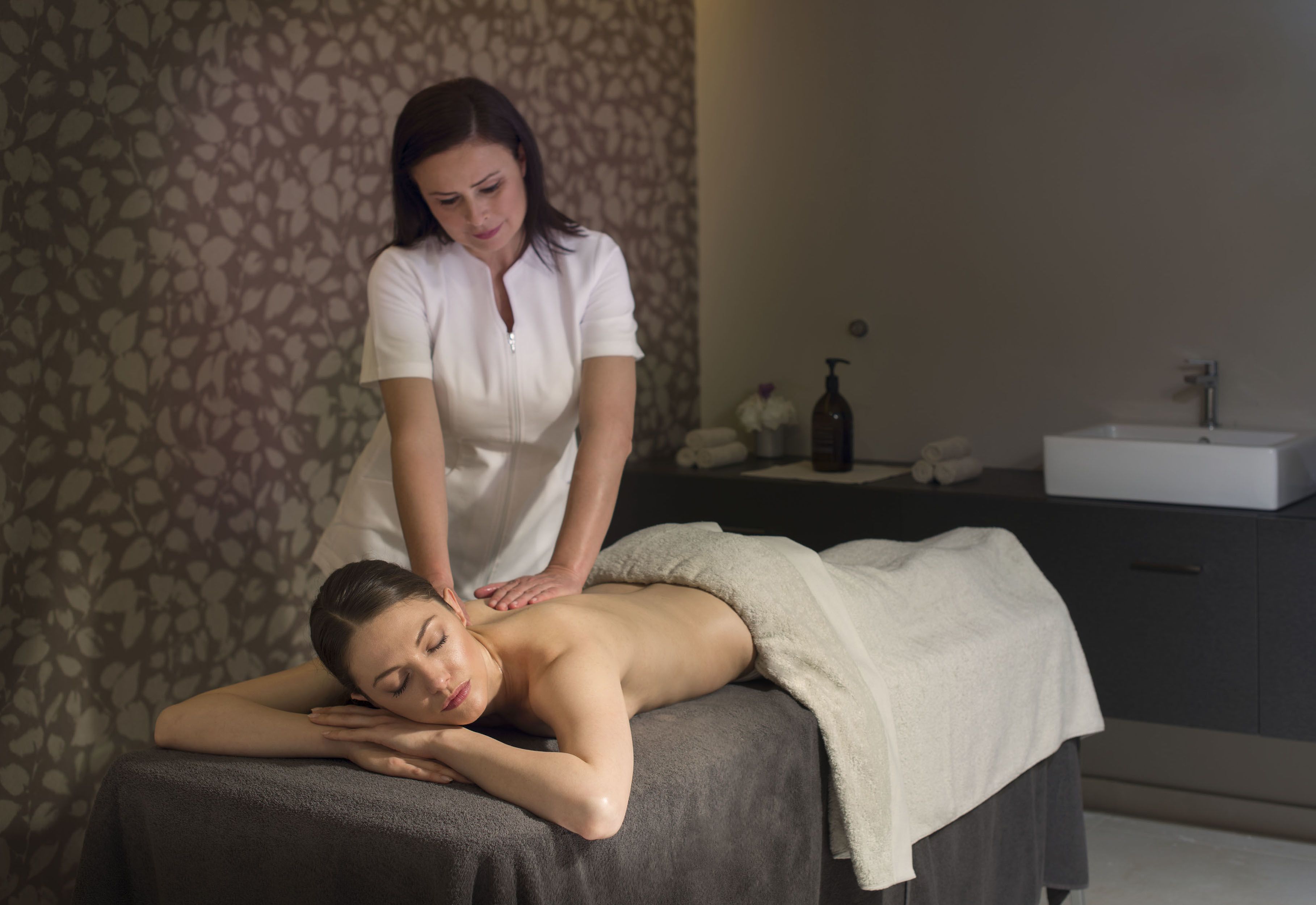 Sessão de massagem em um spa, com parede estampada e pia ao fundo.