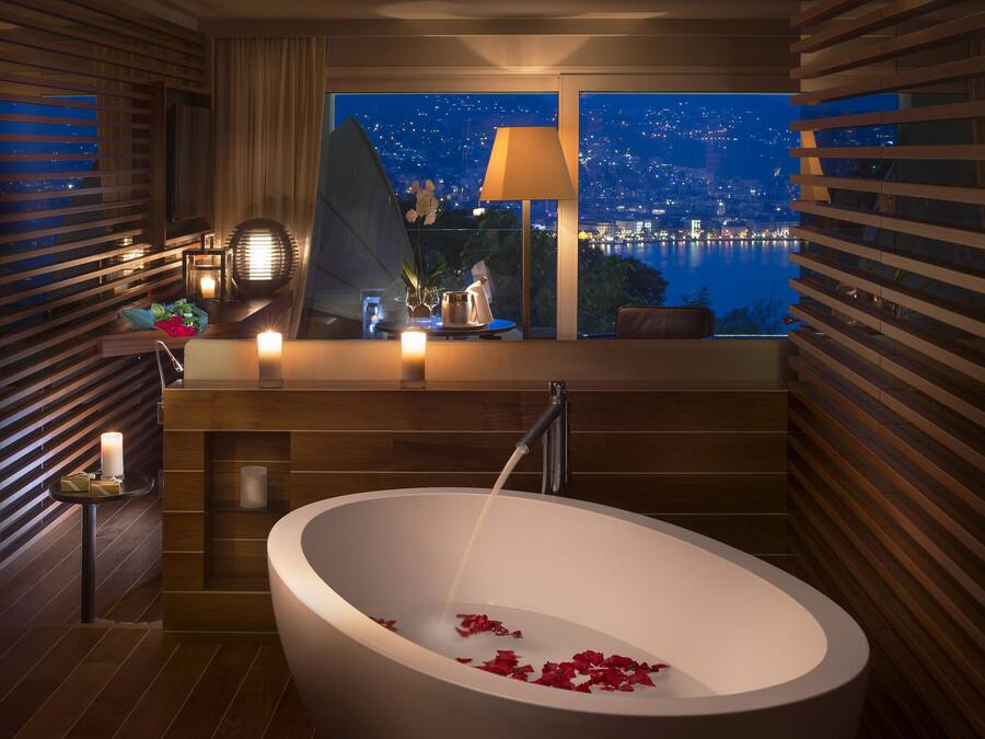 A modern bathroom with a freestanding bathtub, a window with a night view of a cityscape, and a small table with candles and flowers.