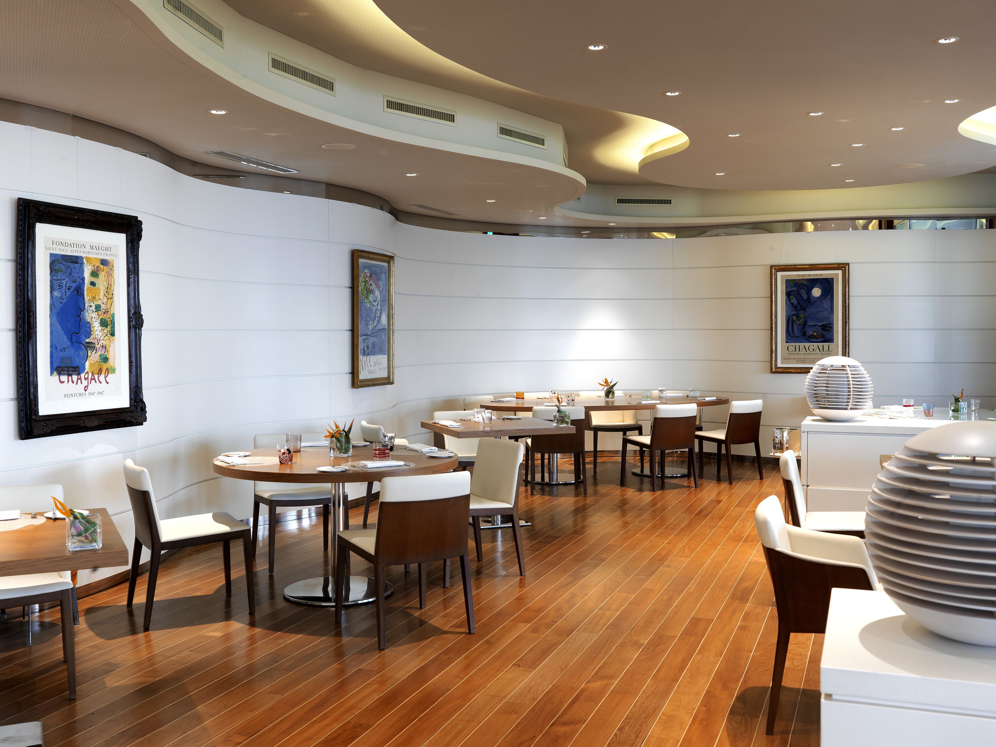 Un espace de restauration moderne avec plusieurs tables et chaises rondes, des murs blancs et des œuvres d’art encadrées.