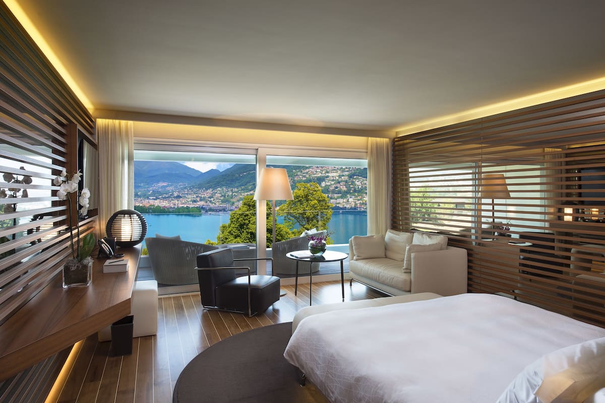 A modern hotel room with a large bed, a sofa, a desk, and a view of a lake and mountains.