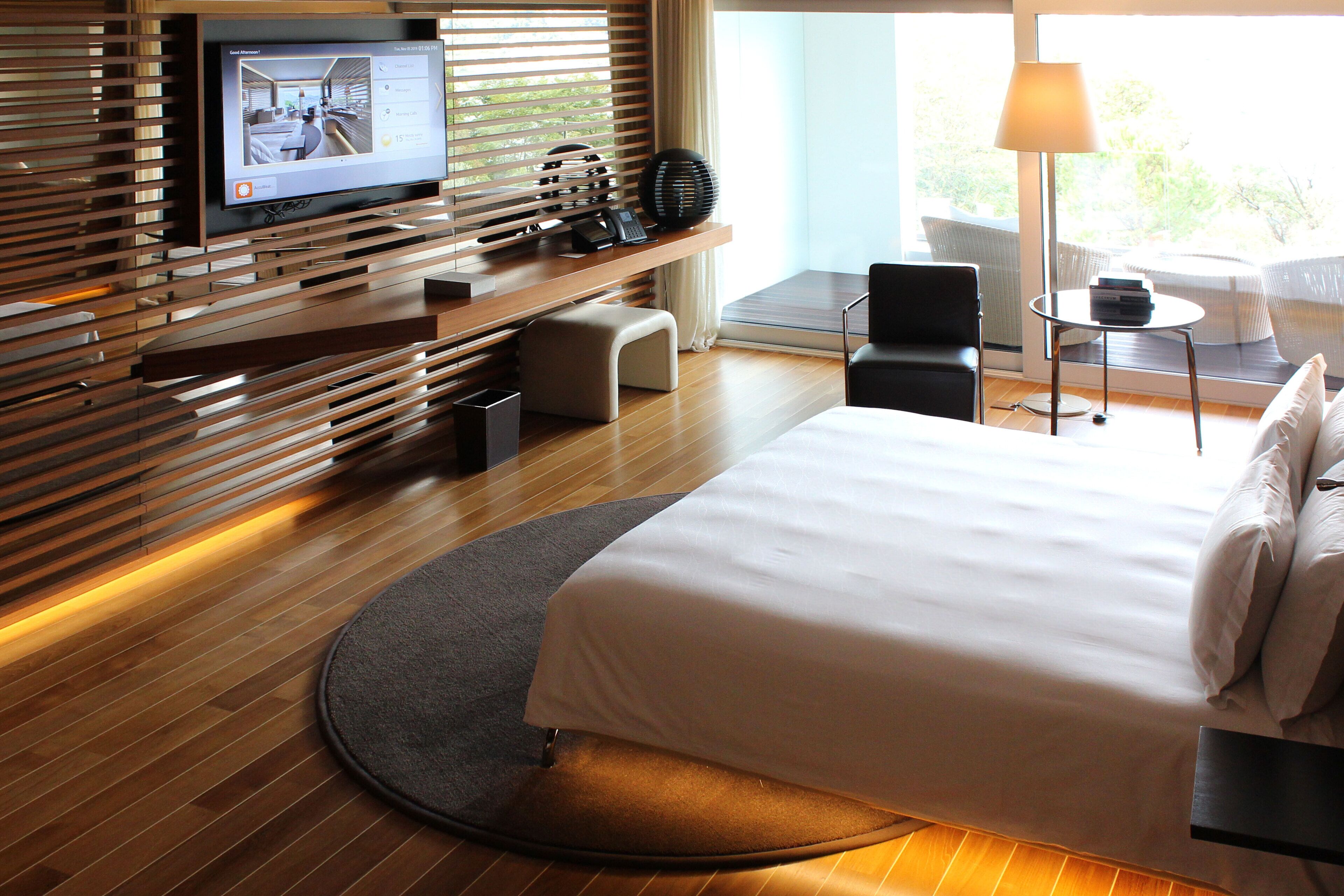 A modern hotel room with a large bed, a television on a wooden shelf, a small table with a lamp, and a view of greenery outside.