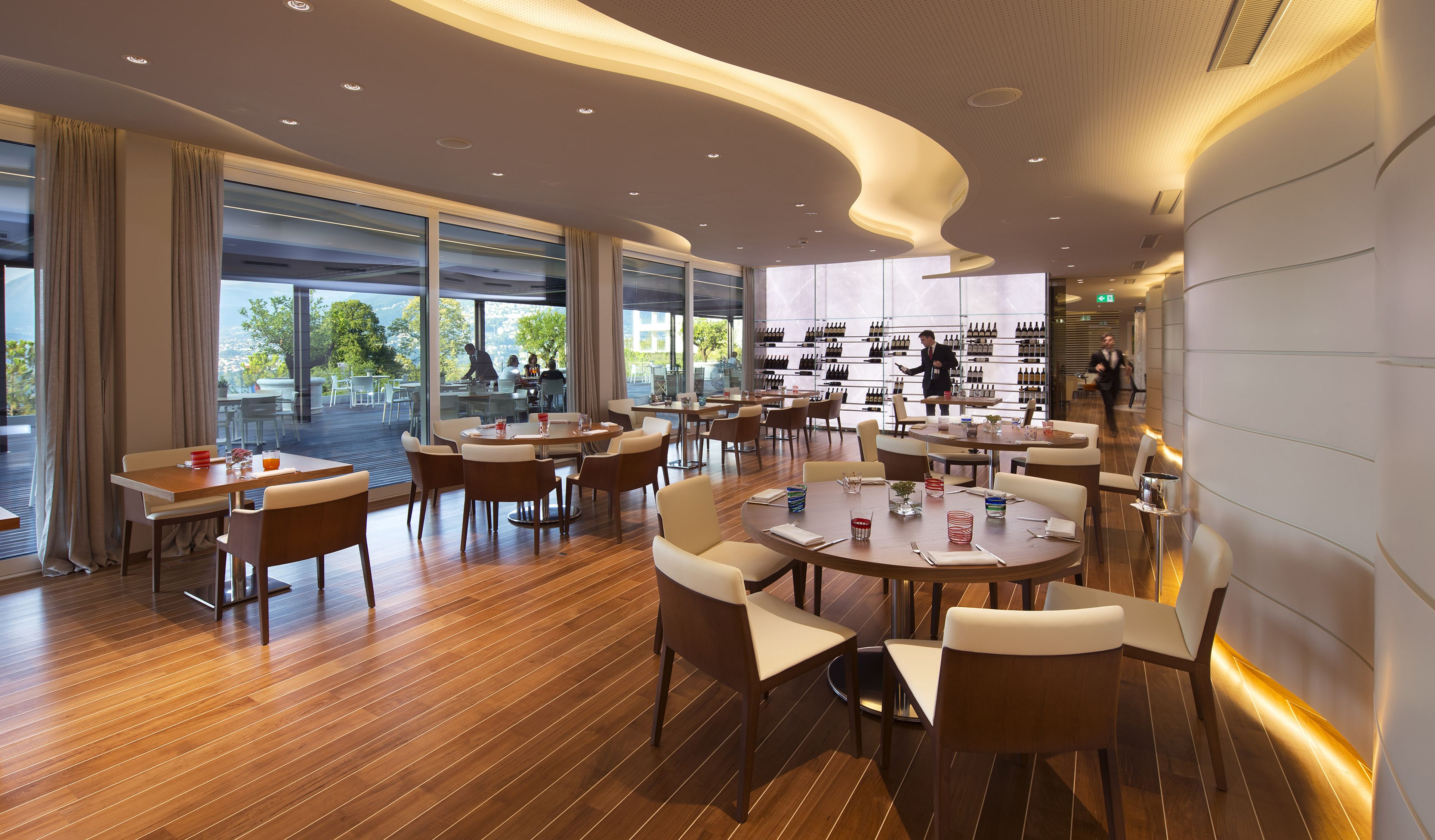 A modern restaurant with wooden flooring, round tables, and chairs. There are large windows with curtains, and a wine display in the background.