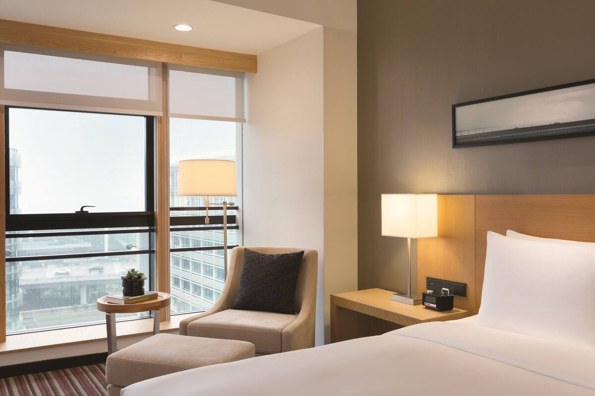 Ein Hotelzimmer mit großem Fenster, einem Bett, einem Sessel, einem kleinen Tisch mit Lampe und einem Nachttisch ebenfalls mit Lampe.