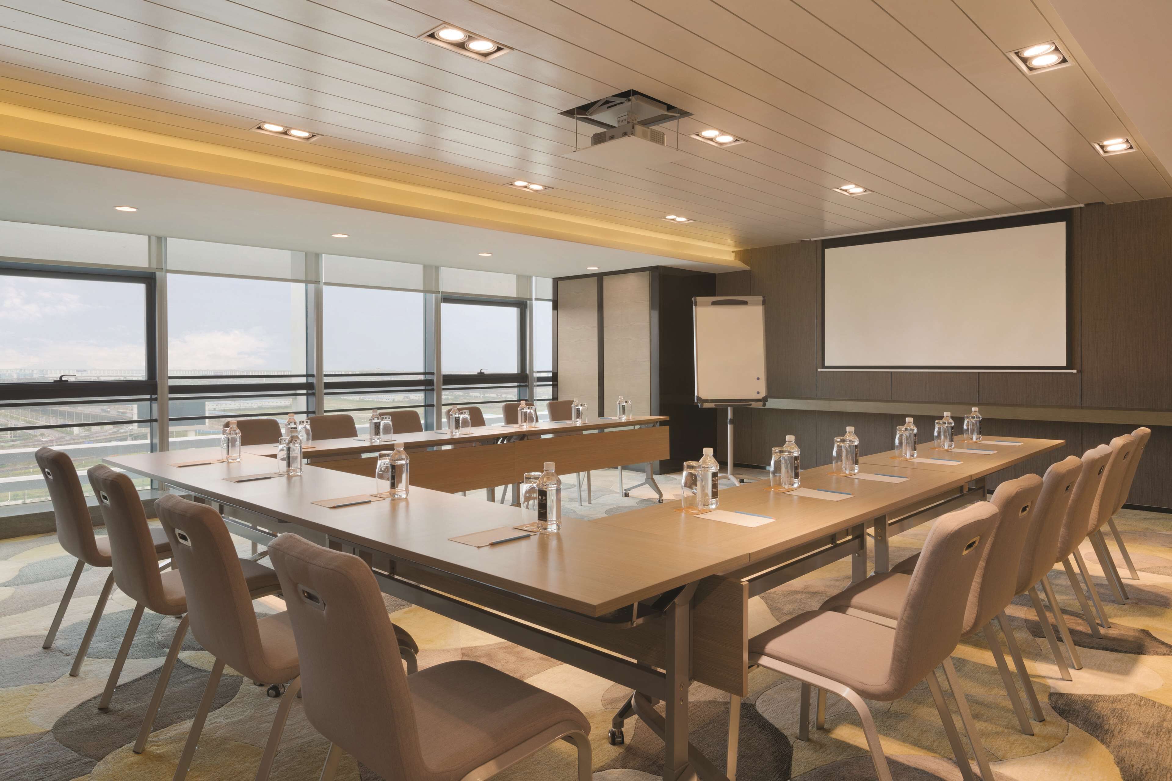 A conference room with a long table, chairs, a projector screen, and a view through large windows.