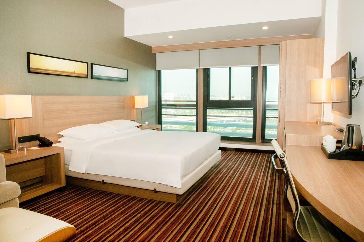 Ein Hotelzimmer mit einem großen Bett, einem Schreibtisch mit Lampe, einem Sessel, einem Fernseher und Blick auf die Stadt durch das Fenster.