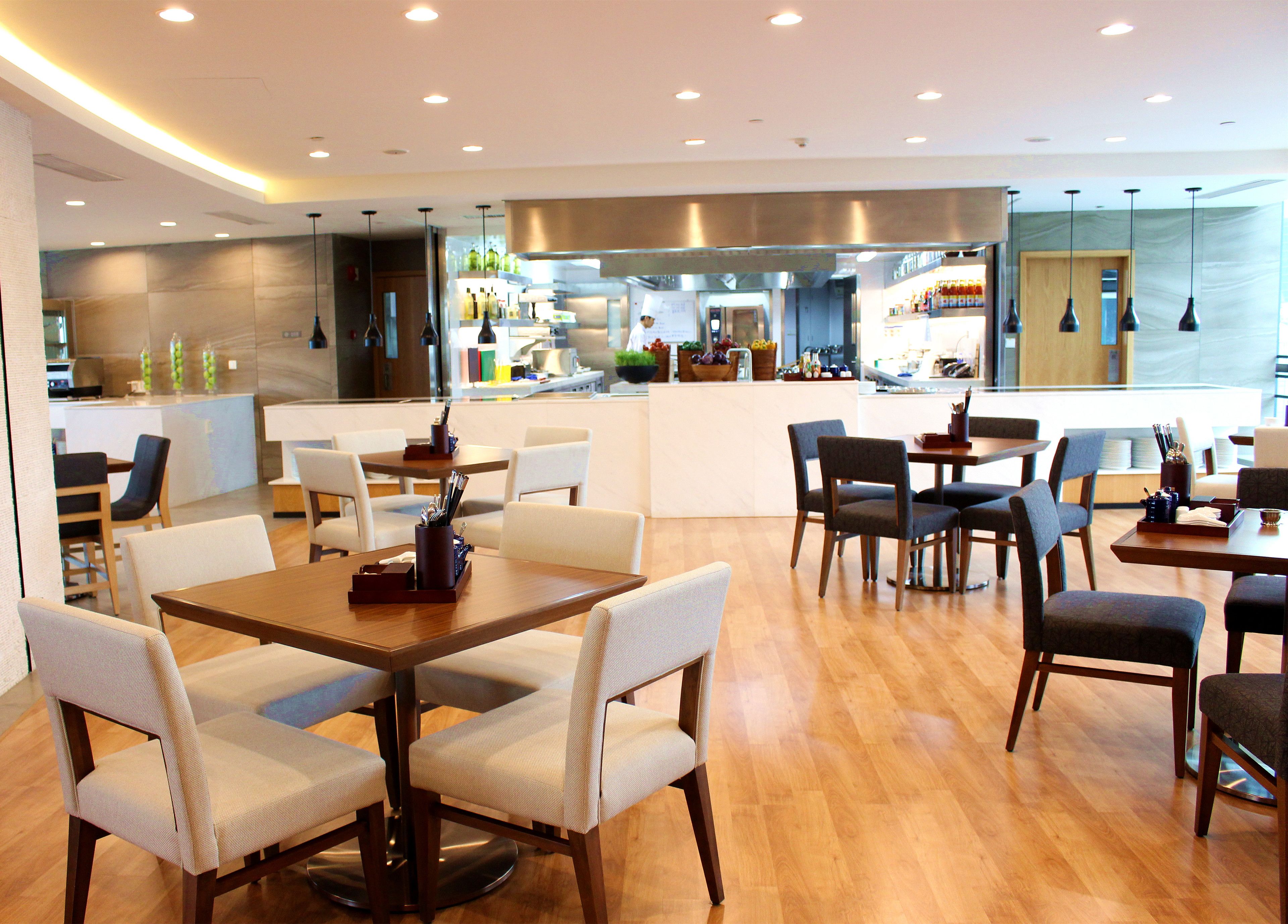 A modern restaurant with wooden tables and chairs, a kitchen in the background, and a bar area.