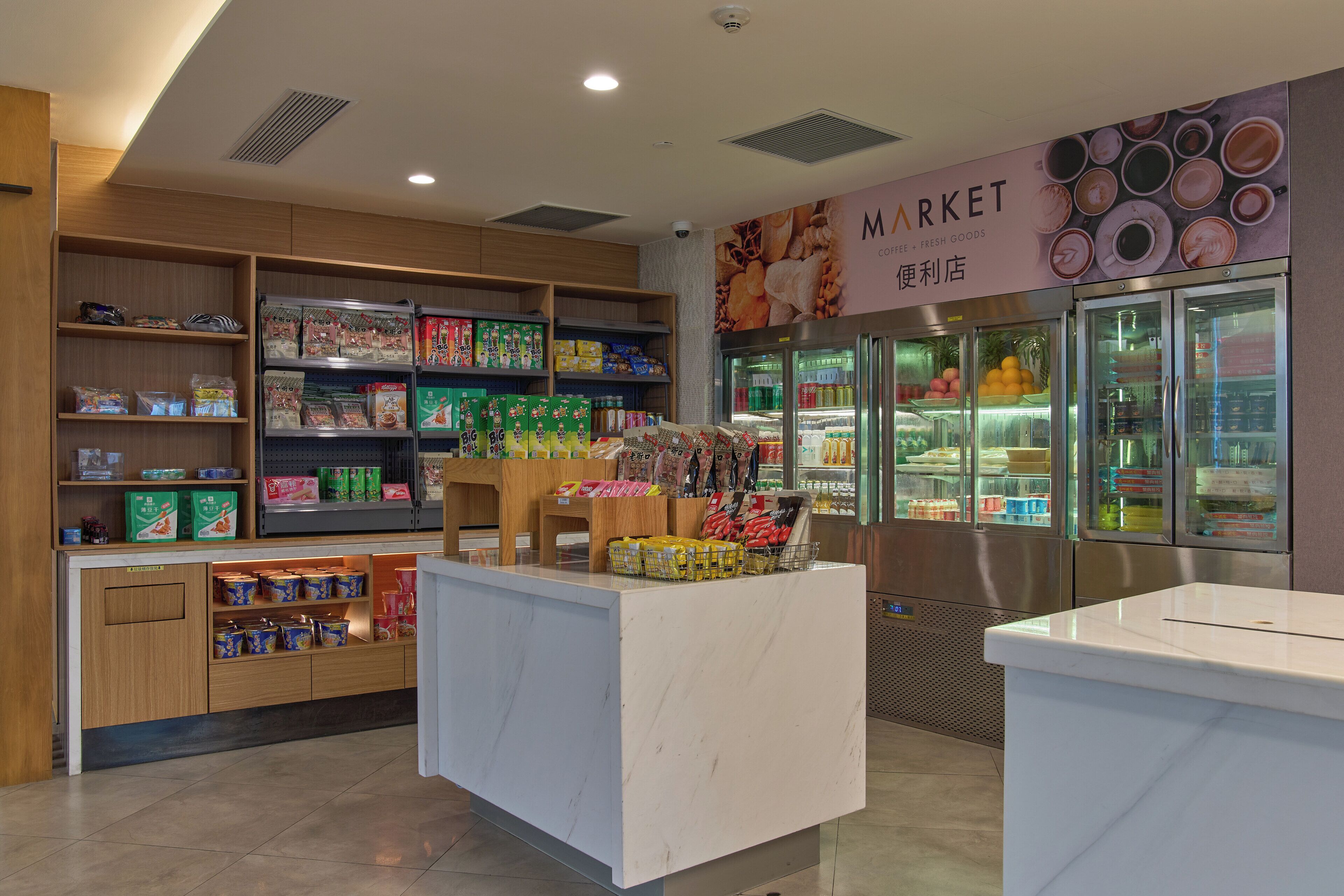 A clean, well-organized market area with shelves stocked with various packaged goods, a refrigerated display case, and a marble counter.
