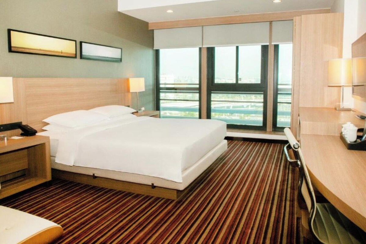 Une chambre d’hôtel dotée d’un grand lit, d’une tête de lit en bois, d’une table de chevet avec une lampe, d’un bureau avec une lampe et offrant une vue sur la ville grâce à de grandes fenêtres.