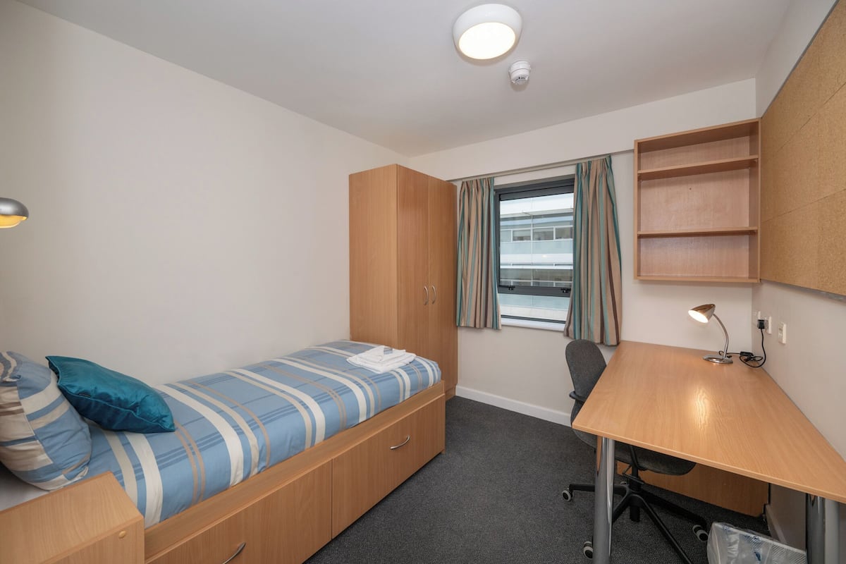 A small, single-room accommodation with a bed, desk, and a window with curtains.