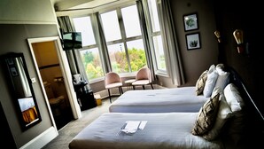 A hotel room with two beds, a TV, a desk, and a window with a view of greenery. - The Brunswick (Shanklin)
