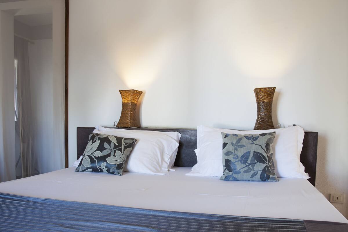 A neatly made bed with white and patterned pillows, two decorative vases on the headboard, and a plain wall in the background.
