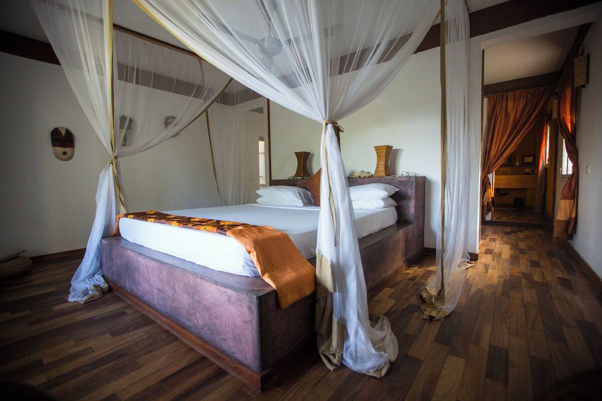 A bedroom with a four-poster bed, wooden flooring, and a view of the hallway.