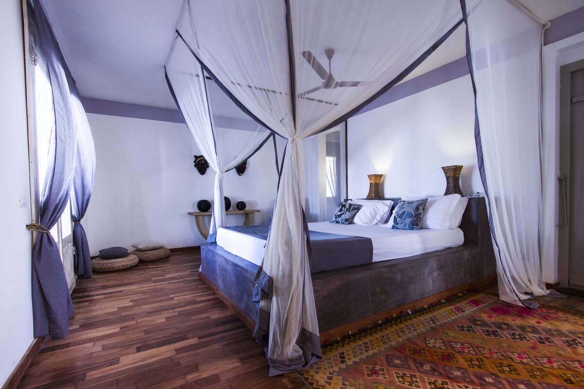 A bedroom with a canopy bed, a ceiling fan, a patterned rug, and a wooden floor.
