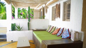 A room with a wooden bench, a small table, and a green bedspread. There are colorful pillows on the bench and a window with a view of greenery outside.