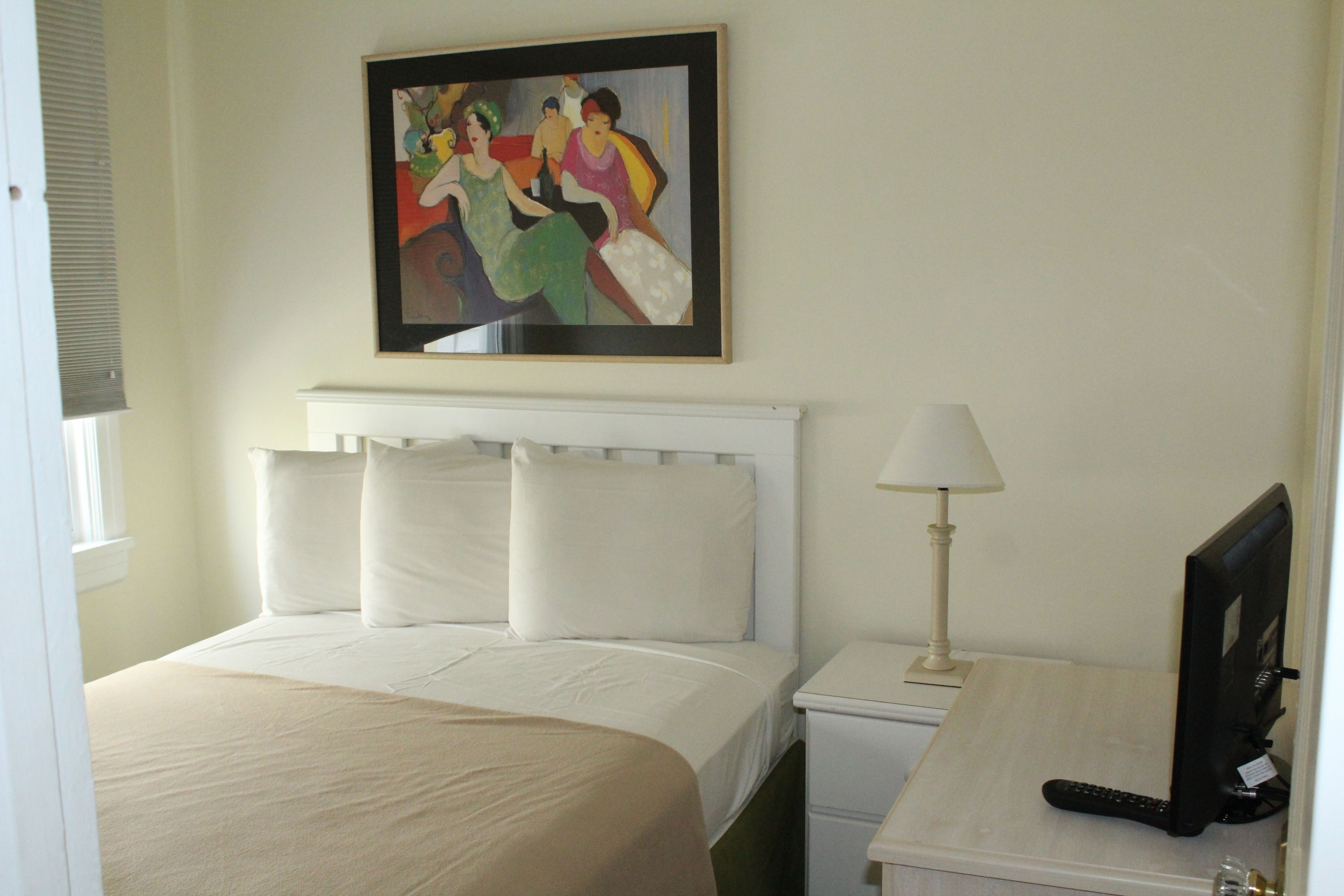 Habitación de hotel con cama, escritorio con lámpara, monitor de computadora y un cuadro en la pared.
