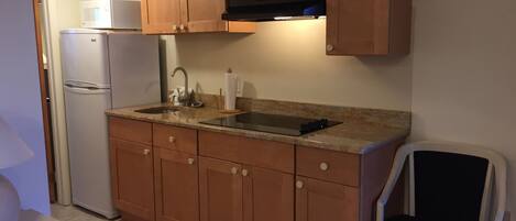 A compact kitchen with wooden cabinets, a built-in microwave, a refrigerator, a sink, and a chair.