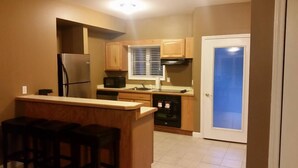 A compact kitchen with a refrigerator, microwave, and stove. There are wooden cabinets and a small island with stools. A door with a frosted glass panel is visible. - Dunmaglas Golf Course (Charlevoix)