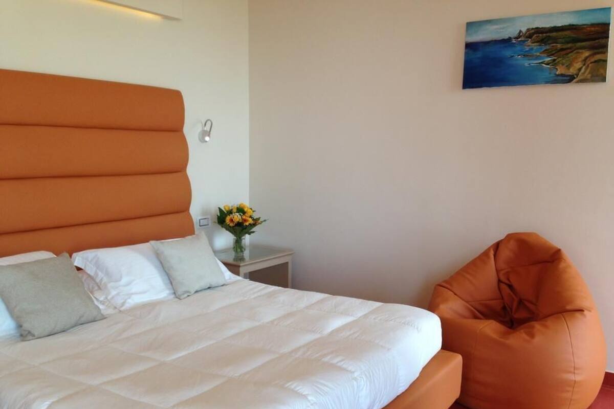 A bedroom with a large bed, an orange bean bag chair, a nightstand with a vase of flowers, and a painting on the wall.