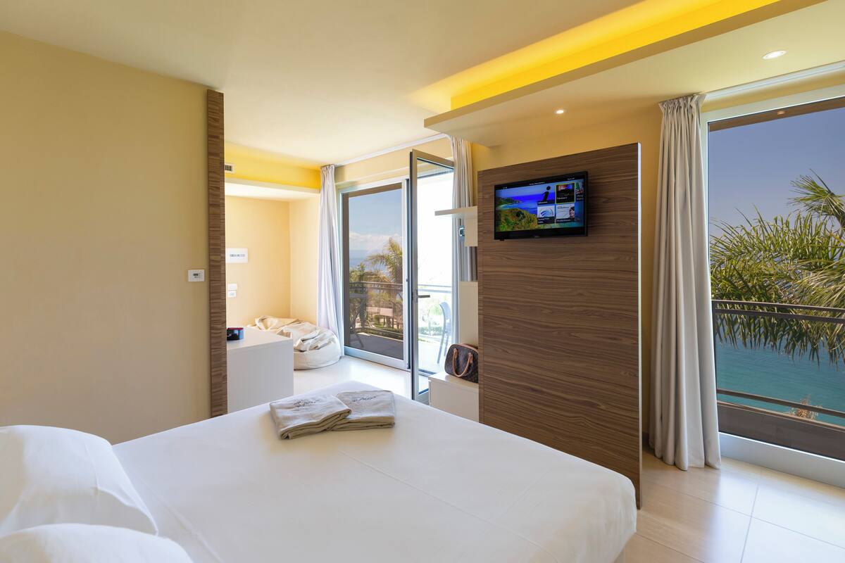 A hotel room with a bed, a television mounted on the wall, a balcony with a view of the sea, and a sliding door leading to a sitting area.