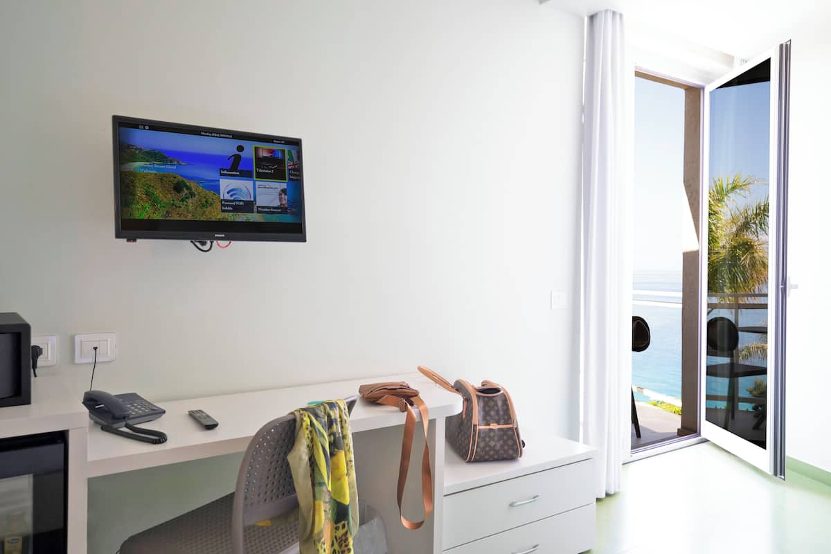 A hotel room with a desk, a television mounted on the wall, a chair, a suitcase, and a view of the ocean through a sliding glass door.