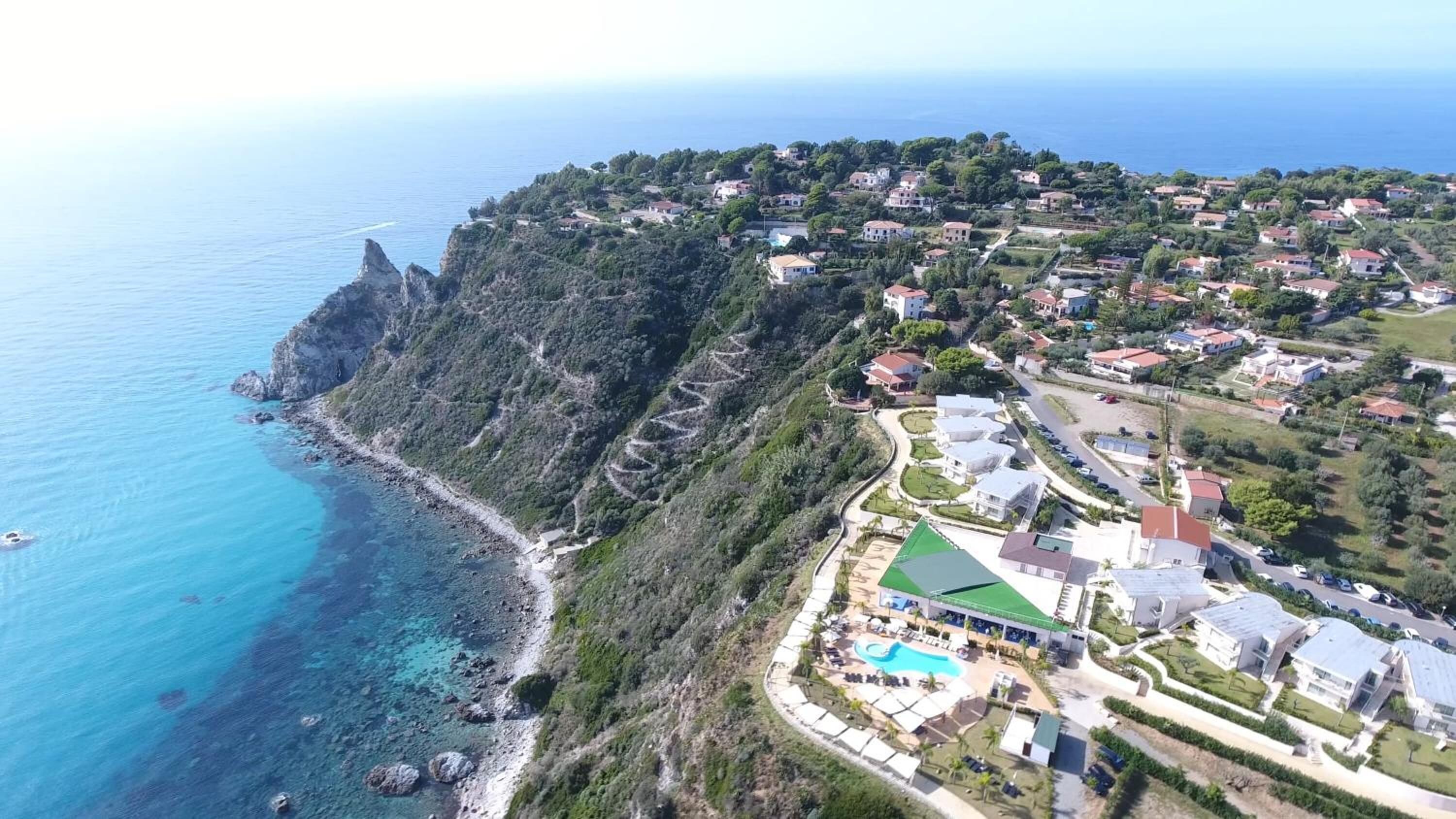 Vue aérienne d’une zone côtière avec un complexe hôtelier comprenant une piscine, plusieurs bâtiments et une côte rocheuse.