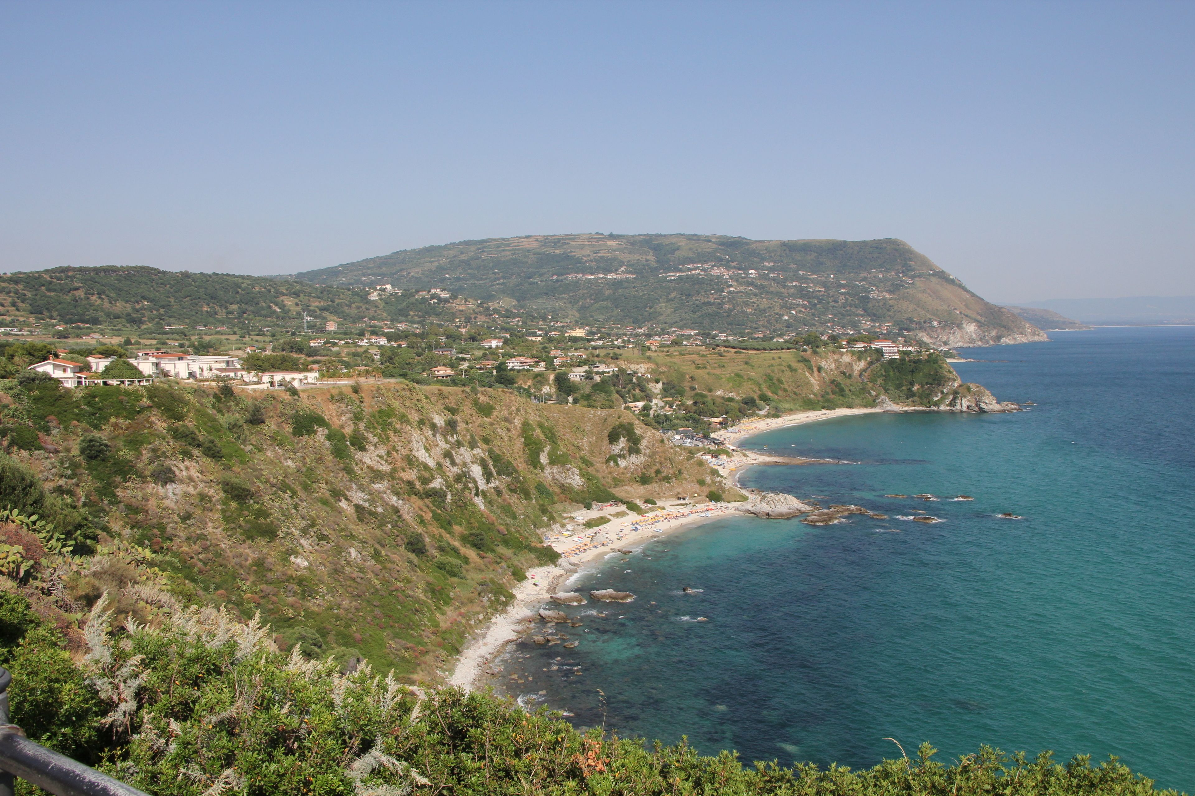 Un paysage côtier avec une plage, une colline parsemée de bâtiments et une mer d’un bleu limpide.