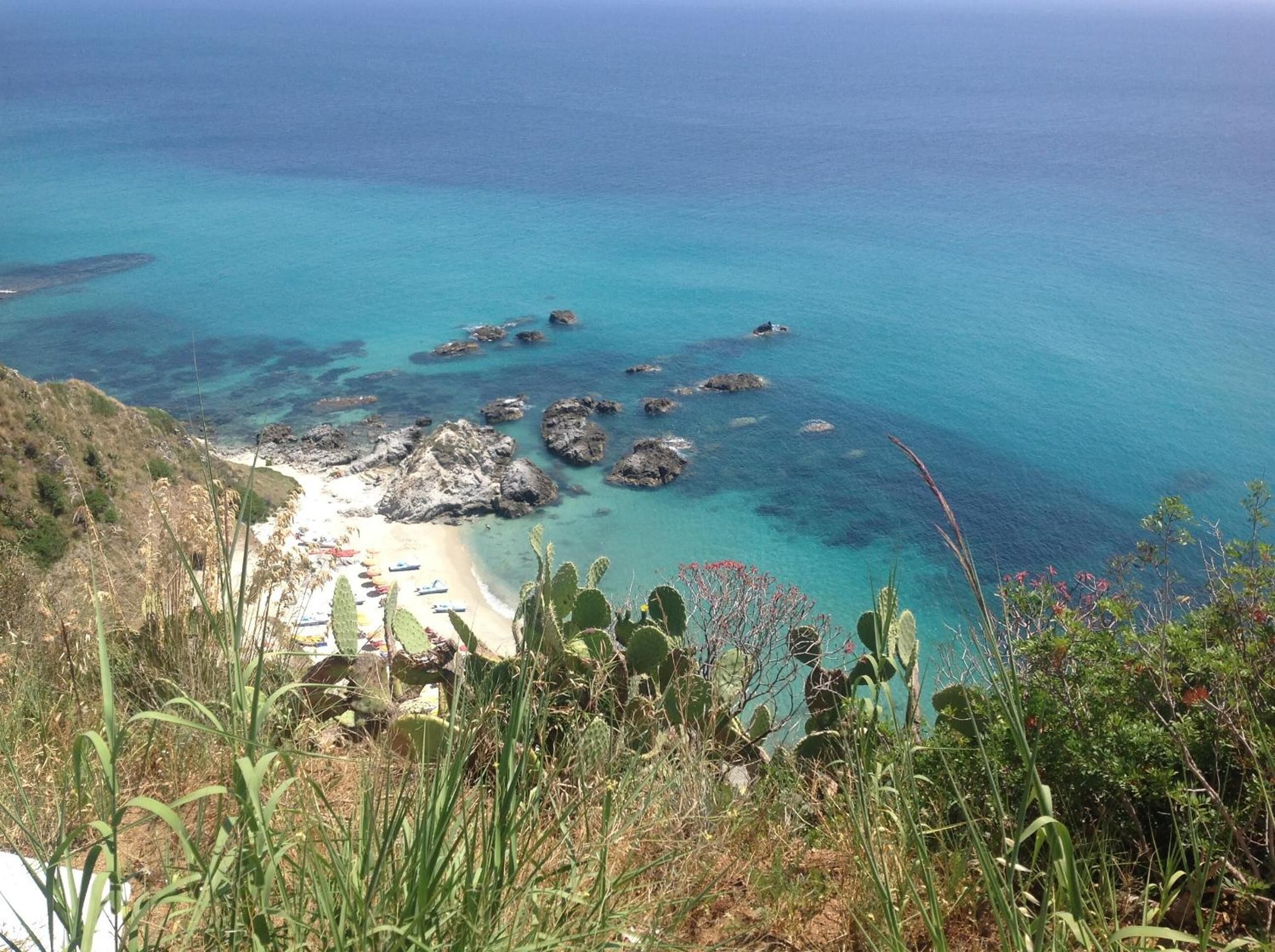 Paesaggio costiero con acque turchesi cristalline, affioramenti rocciosi e una spiaggia sabbiosa.