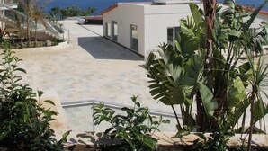 Un edificio moderno con techo plano y ventanas amplias, rodeado de plantas tropicales y vistas a un cielo azul despejado y al océano.