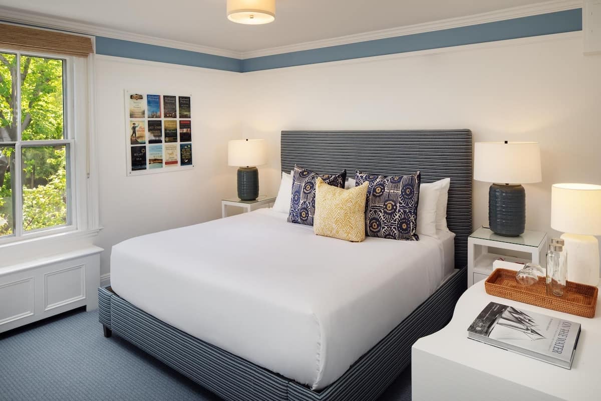 Um quarto bem arrumado com uma cama grande, duas mesas de cabeceira com luminárias, um painel de livros na parede e uma janela com vista para vegetação.
