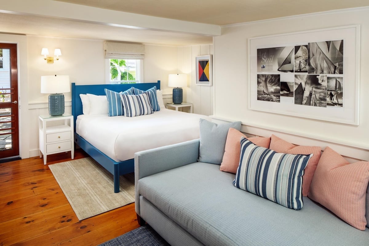 Quarto de hotel com cama, sofá, mesa de cabeceira com abajur e porta-retrato fixado na parede.