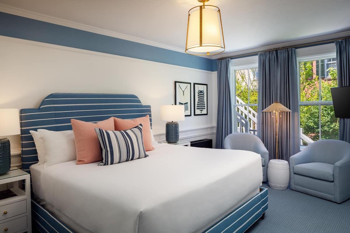 Quarto de hotel moderno com uma cama grande, área de estar com uma poltrona e apoio para os pés, mesa de cabeceira e uma janela ampla com cortinas.