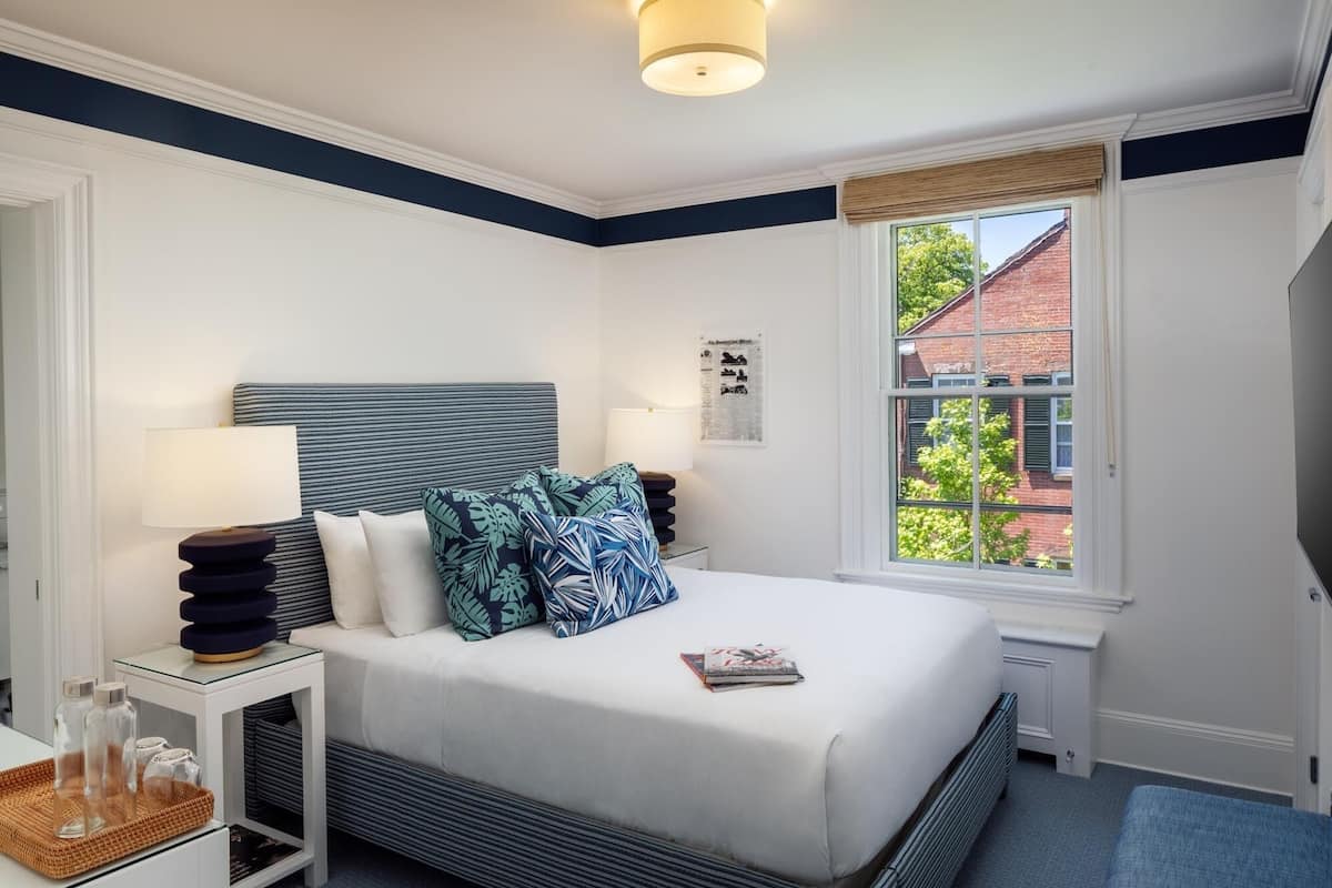 Quarto bem arrumado com cama, mesas de cabeceira, janela com vista para um edifício de tijolos e uma luminária fixada na parede.