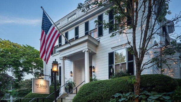 A two-story hotel with a white facade, a flagpole with an American flag, and a sign reading "76 Main Ink Press Hotel".