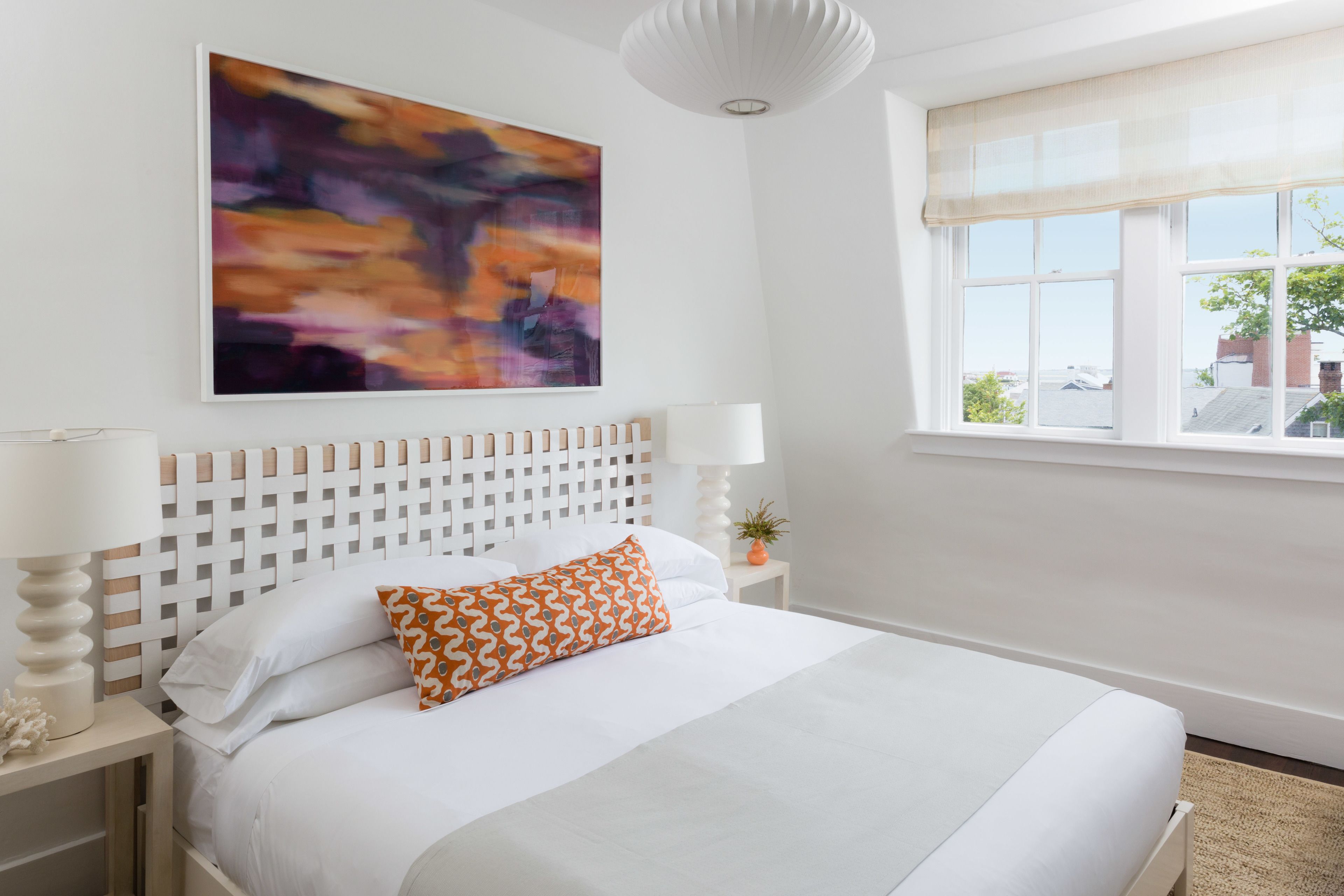 A neatly made bed with white linens and an orange patterned pillow, a white headboard, a white nightstand with a lamp, a wall-mounted abstract painting, and a window with a view of buildings and trees.