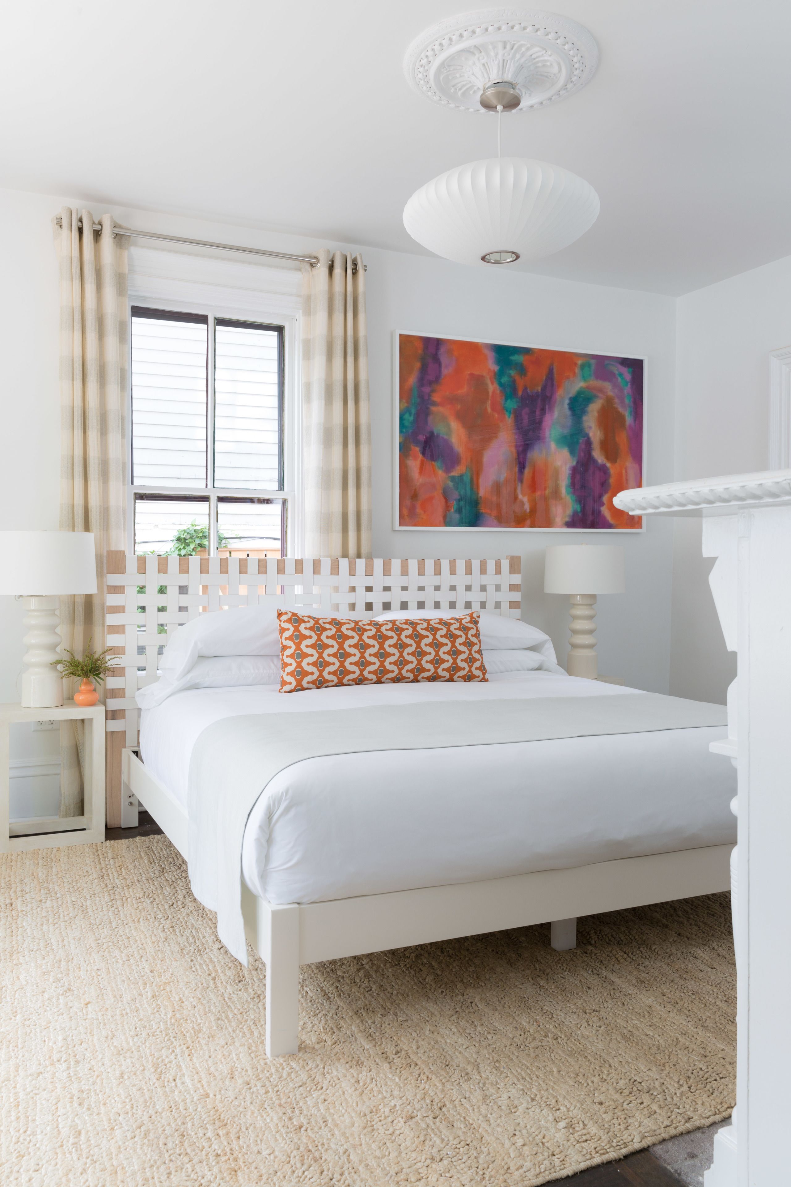 A bedroom with a white bed, beige curtains, a colorful abstract painting, and a white ceiling fixture.