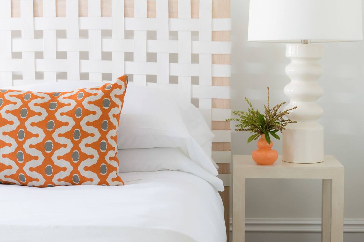 A bedroom with a white bed, an orange patterned pillow, a small side table with a lamp and a plant, and a colorful abstract painting on the wall.