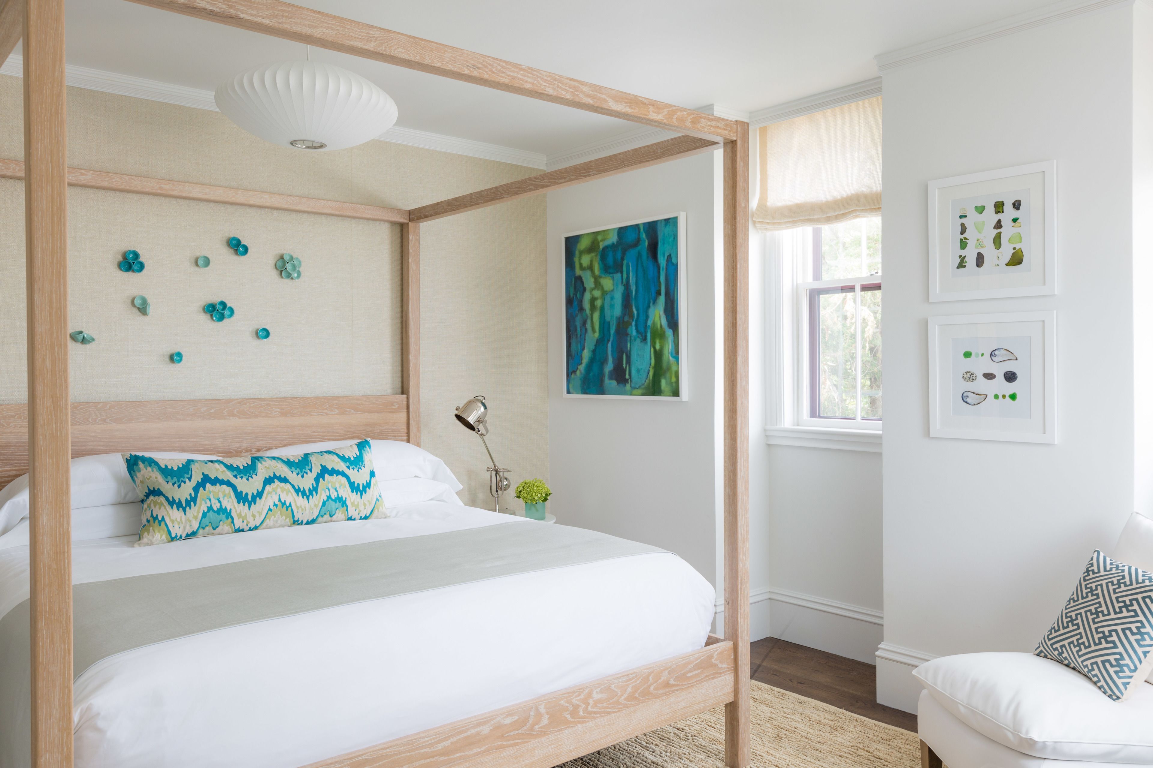 A four-poster bed with a white and blue patterned pillow, a modern abstract painting, and framed botanical prints on the wall.