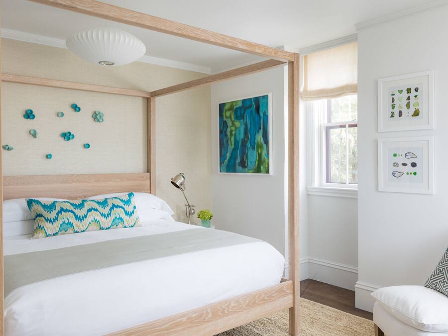 A four-poster bed with a white and blue patterned pillow, a modern abstract painting, and framed botanical prints on the wall.