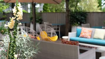 Un patio al aire libre con mobiliario de mimbre, un sofá con cojines y una planta alta con flores blancas.