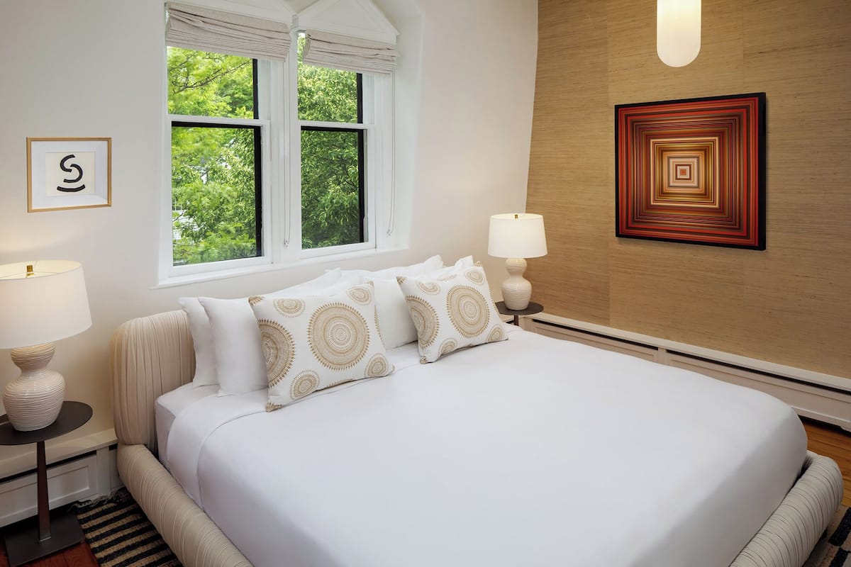 A neatly arranged bedroom with a large bed, two bedside lamps, a framed artwork, a window with a view of greenery, and a geometric wall art piece.