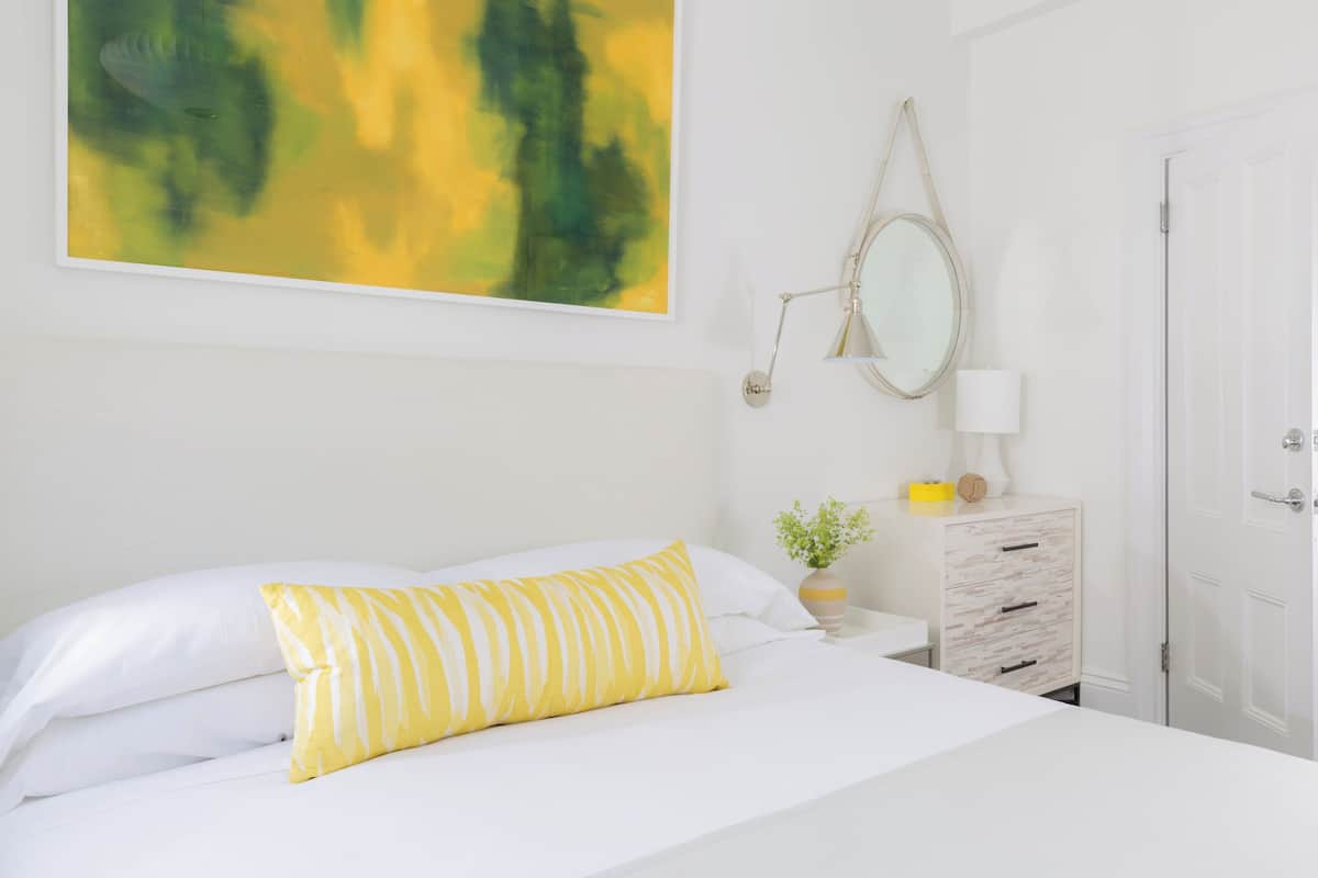 A neatly made bed with white and yellow striped pillows, a bedside table with a lamp, a small plant, and a white dresser with drawers.