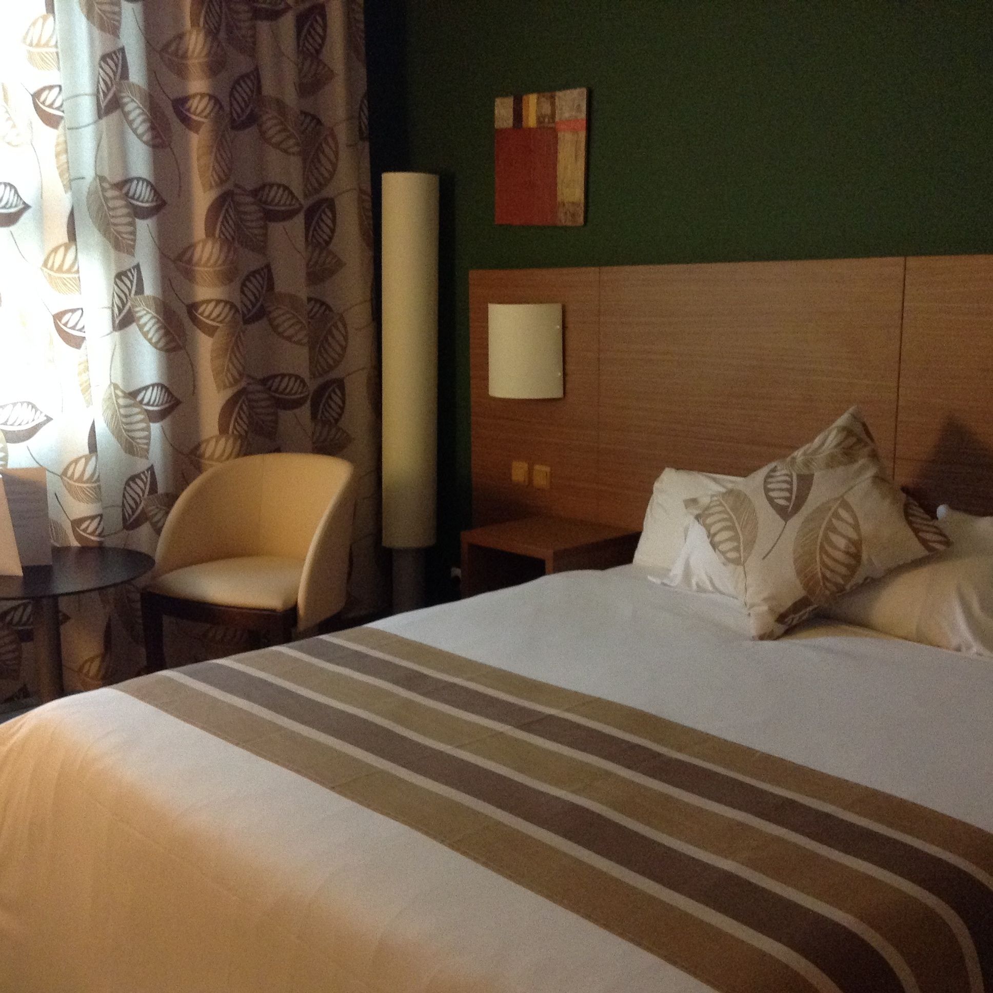 Una habitación de hotel con cama, una silla, una mesita de noche, una lámpara y un cuadro en la pared.