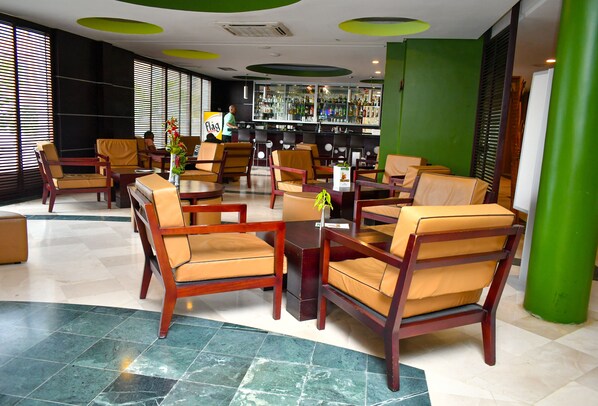 A lobby with wooden chairs and tables, a bar area with bottles, and a green accent wall.