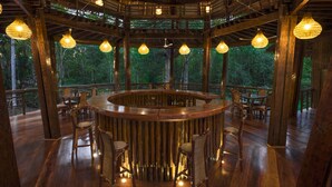 A wooden bar area with a circular counter, surrounded by wicker chairs, and large hanging lights. - Treehouse Lodge (Saquena)