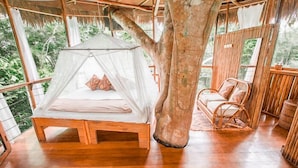 A treehouse bedroom with a bed under a canopy, a wooden floor, and a wicker chair. - Treehouse Lodge (Saquena)