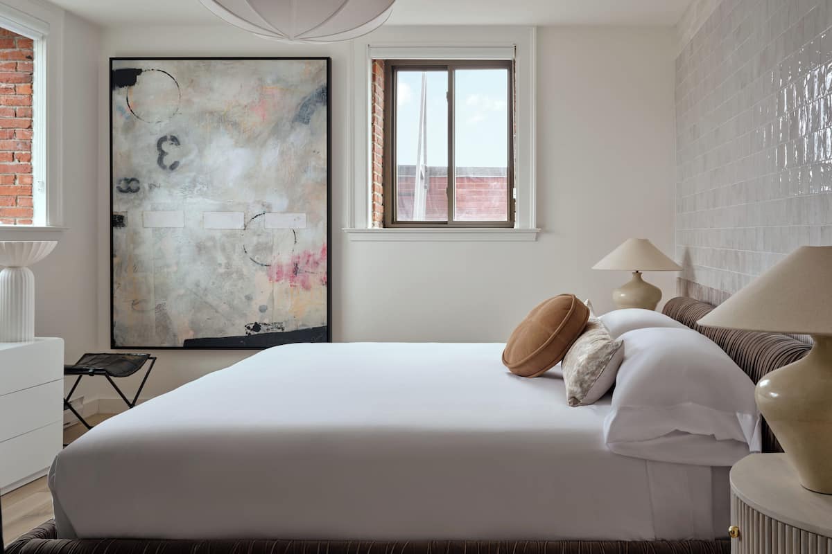A modern bedroom with a large bed, a view of the city through a window, and a large abstract painting on the wall.